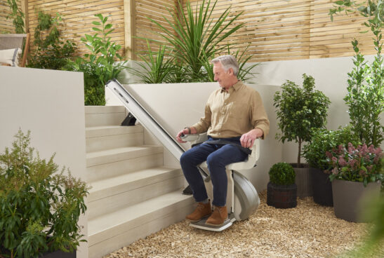 A man rides a stair lift outdoors, ascending a short flight of steps surrounded by potted plants and greenery in a garden setting.