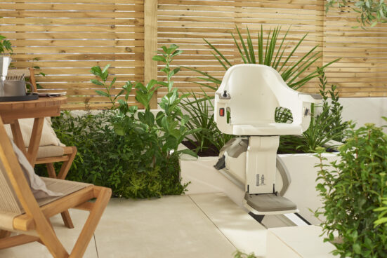 A white stairlift is installed outdoors on a patio surrounded by green plants and wooden furniture, with a wooden privacy fence in the background.