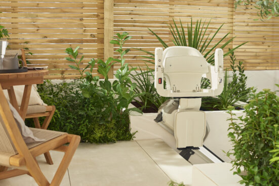 A stairlift chair positioned on a rail next to a cozy outdoor patio with wooden furniture, surrounded by green plants and a wooden privacy fence in the background.