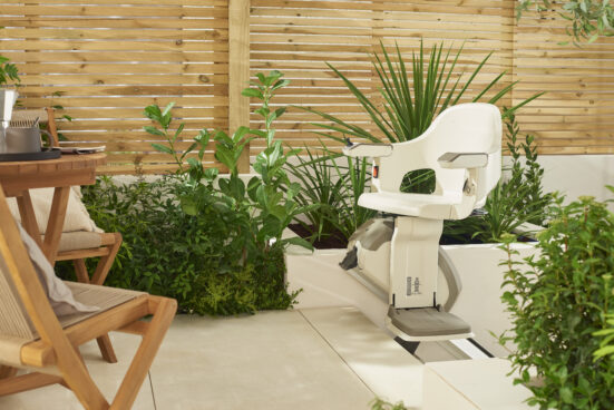 A modern stairlift is installed outdoors next to green plants and wooden patio furniture, with a wooden privacy fence in the background. The setting appears bright and inviting.