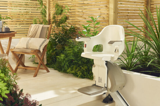 A modern stairlift chair is installed along a railing in a bright, plant-filled patio with wooden privacy panels, a wooden chair, and a small table with a teapot and cushion nearby.