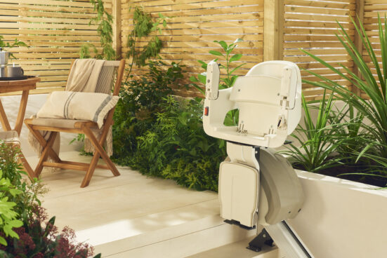 A beige stairlift is installed along outdoor steps next to a wooden privacy screen, green plants, and a wooden chair with a folded blanket on a patio.