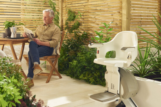 A man sits on a wooden chair reading a newspaper in a bright, plant-filled patio. Nearby, a modern stair lift is installed along the steps, surrounded by lush greenery and wooden fencing.