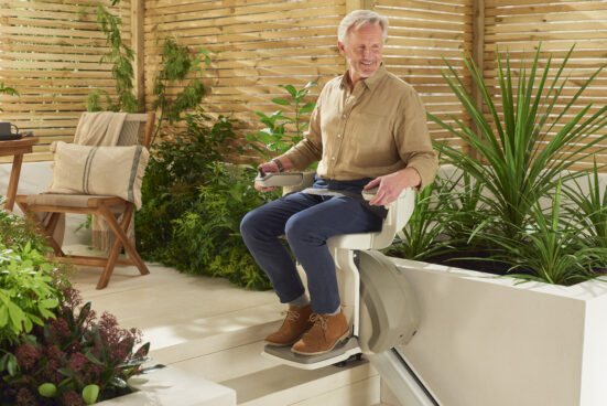 An older man sits on an outdoor stairlift surrounded by green plants and wooden patio furniture, smiling and looking to the side on a sunny day.