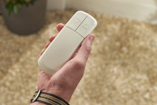 A person wearing a watch holds a white remote control with two triangular buttons, one pointing up and one pointing down, against a beige, textured background.