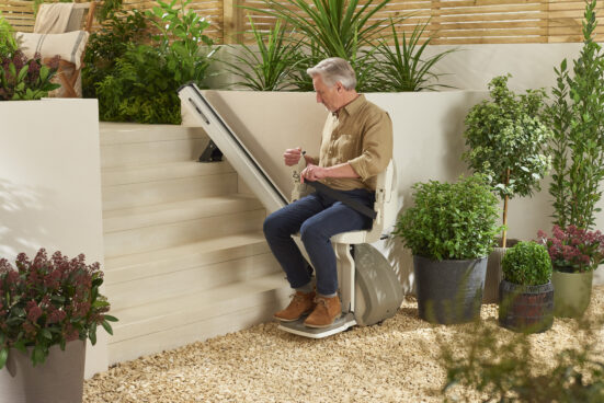 An older man sits on a stairlift at the base of outdoor steps, surrounded by potted plants and greenery, preparing to ascend the stairs in a well-landscaped garden area.
