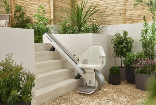 A stairlift is installed alongside a small indoor staircase surrounded by lush green plants, modern planters, and light-colored stone walls in a bright, garden-like space.