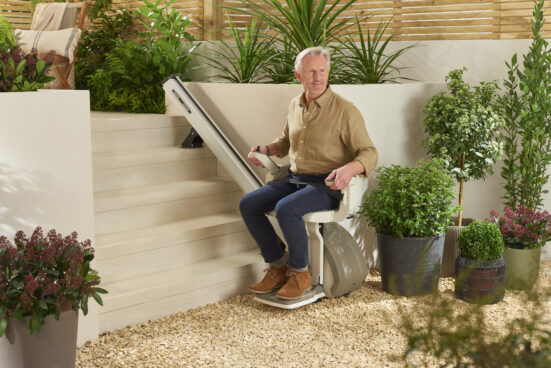 An older man sits on a stairlift at the bottom of outdoor steps, surrounded by potted plants and greenery in a garden setting, smiling and holding the lift’s controls.