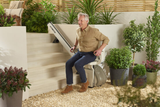 An older man sits on a stairlift at the bottom of outdoor steps, surrounded by green plants and potted shrubs in a garden setting. He is smiling and holding the stairlift control.