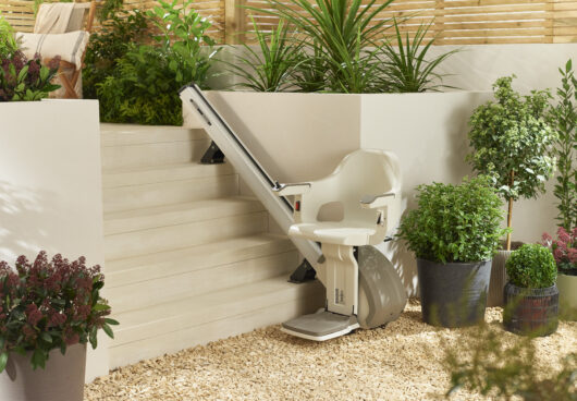 A stairlift chair is installed on a set of white outdoor stairs surrounded by green plants and potted flowers on a pebble-covered patio.