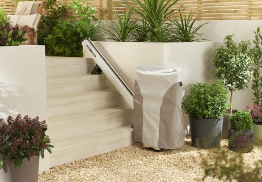 A stair lift with a protective cover is installed alongside white outdoor steps, surrounded by various green potted plants in a landscaped garden area with a pebble ground.