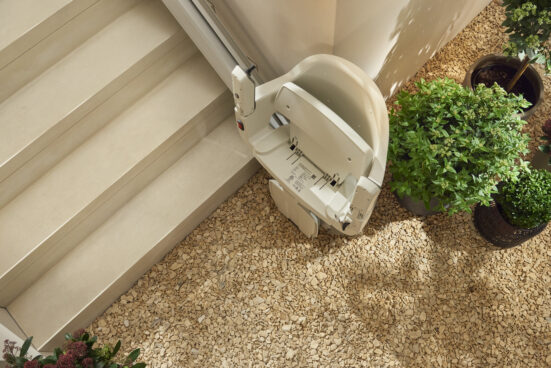 A stairlift chair is positioned at the base of light-colored indoor stairs, surrounded by potted green plants on a pebble-covered floor.