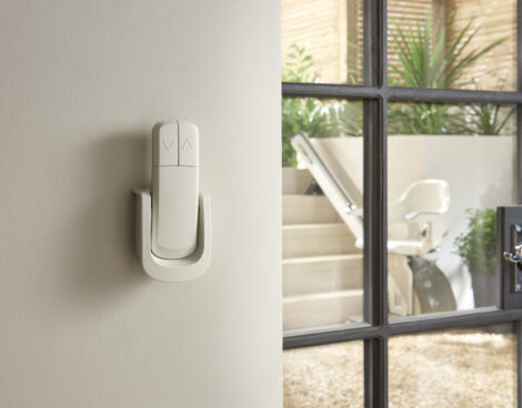 A stairlift control switch is mounted on a cream-colored wall near a window. Through the window, a stairlift chair is visible on a staircase next to indoor plants.