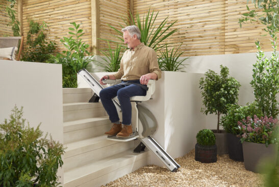 An older man sits on a stairlift, smiling as he descends outdoor steps in a garden with lush green plants, wooden fence, and white stone ground.