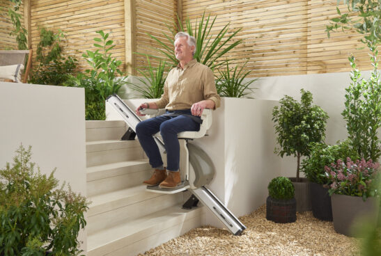 An older man sits on a stairlift going up outdoor steps in a garden, surrounded by green plants and wooden fencing.