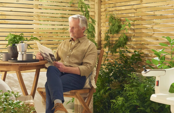 An older man sits on a wooden chair in a sunlit garden patio, reading a newspaper. A small table with a coffee maker is beside him, and green plants surround the area, giving a peaceful, relaxed atmosphere.