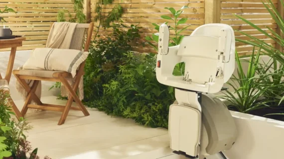 A HomeGlide Outdoor Stair Lift is installed on an outdoor patio next to green plants and wooden seating, with wooden privacy panels and a folded blanket on a chair in the background.