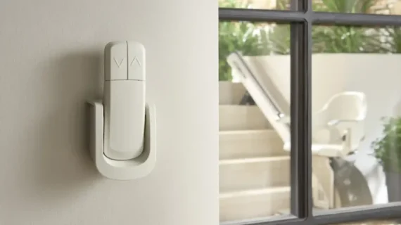 Wall-mounted remote control with up and down buttons for the HomeGlide Outdoor Stair Lift, shown next to a window with the stair lift visible on a set of indoor stairs outside.