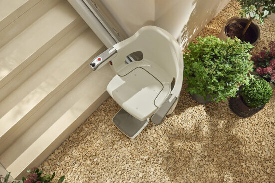 A stairlift chair is installed next to a set of indoor stairs, surrounded by potted green plants and a floor covered with small pebbles.
