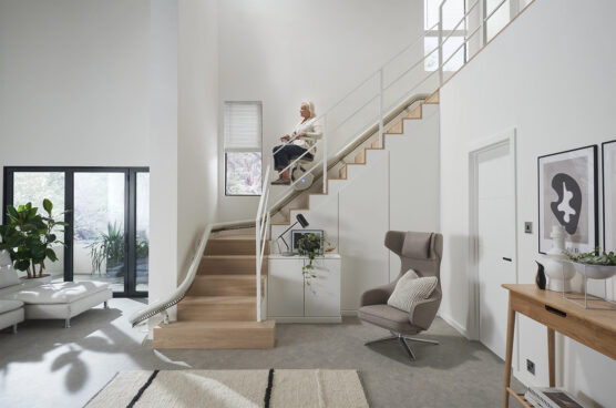 A woman sits on a stairlift going down a wooden staircase in a modern, bright living room with large windows, plants, and contemporary furniture.