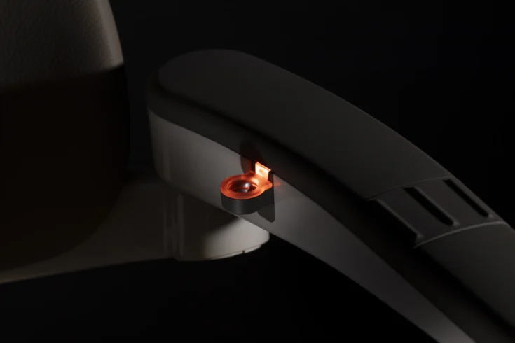 A close-up of a dark-colored chair armrest showcases its Light Assist feature, with a small orange light illuminating a circular button on its side, set against a dark background.