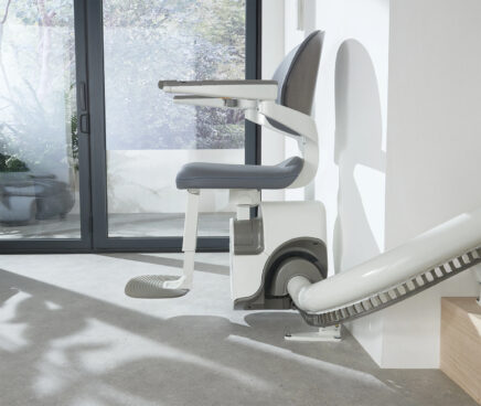 A modern stairlift chair with a gray seat and backrest is positioned on a rail by a staircase in a bright room with large glass doors and greenery visible outside.