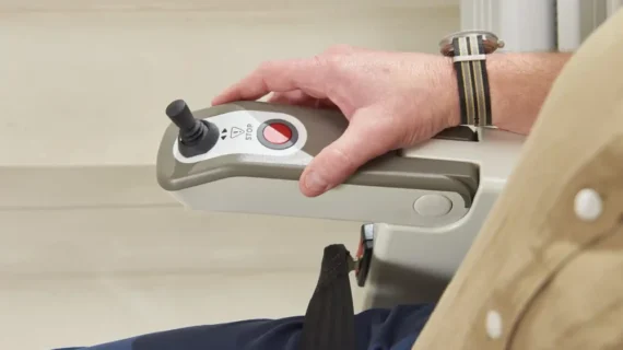 A close-up of a person's hand operating the control panel of a HomeGlide Outdoor Stair Lift, showing a joystick and a red stop button. The person is wearing a striped wristwatch.