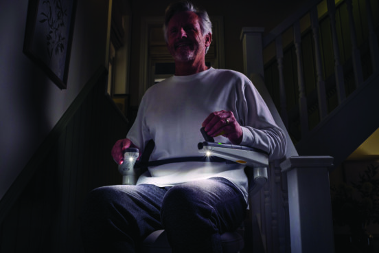 An older man sits on a stairlift with Light Assist, built-in lights glowing from the armrests and illuminating the dim hallway. He wears a white long-sleeve shirt and gray pants, with stair railing and framed art in the background.