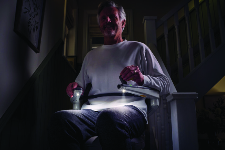 An older man sits on a stairlift with Light Assist, built-in lights glowing from the armrests and illuminating the dim hallway. He wears a white long-sleeve shirt and gray pants, with stair railing and framed art in the background.