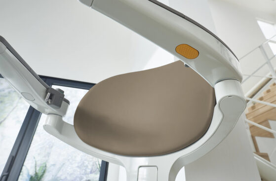 Close-up view of a modern stairlift chair with a beige seat and white frame, installed on a staircase with a window and natural light in the background.
