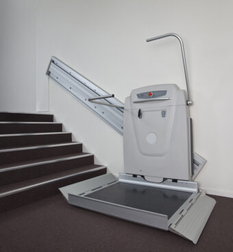 A stairlift platform for wheelchair users, part of the Platform Lifts range, is installed beside a staircase to enable accessible movement between floors. The platform is folded down and ready for use, with rails mounted along the stairs.
