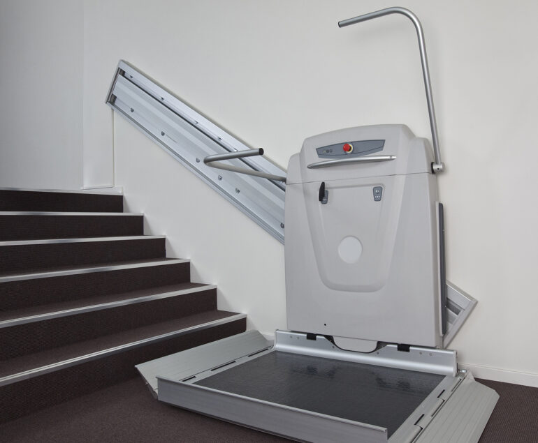 A stairlift platform for wheelchair users, part of the Platform Lifts range, is installed beside a staircase to enable accessible movement between floors. The platform is folded down and ready for use, with rails mounted along the stairs.