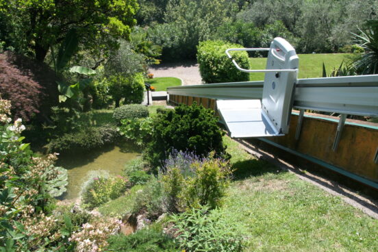 A motorized outdoor stair lift for wheelchairs, part of the Platform Lifts range, descends along a rail through a lush, green garden with trees, shrubs, and a small pond, leading to a gravel path below.