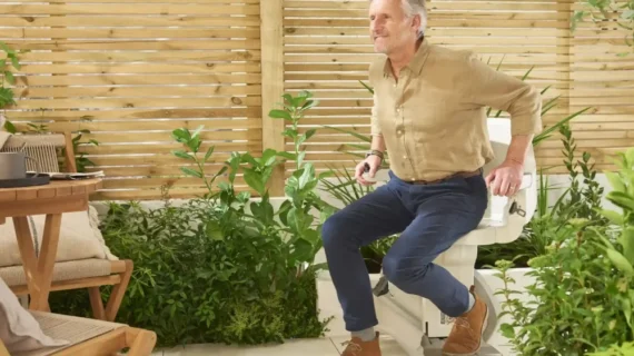 An older man in casual clothes smiles while standing up from a toilet in a bright, leafy outdoor patio area with wooden fencing, green plants, and a nearby HomeGlide Outdoor Stair Lift blending into the scenery.