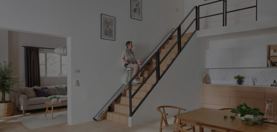 A person using a stairlift to ascend a wooden staircase in a modern, bright home with a kitchen and dining area in the foreground and a living room visible in the background.