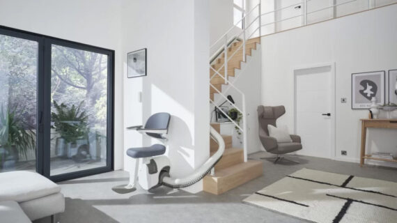 Modern indoor stair lift installed on a curved staircase in a bright contemporary home, featuring a cushioned seat, white rail system, and open-plan living area with large windows and natural light.