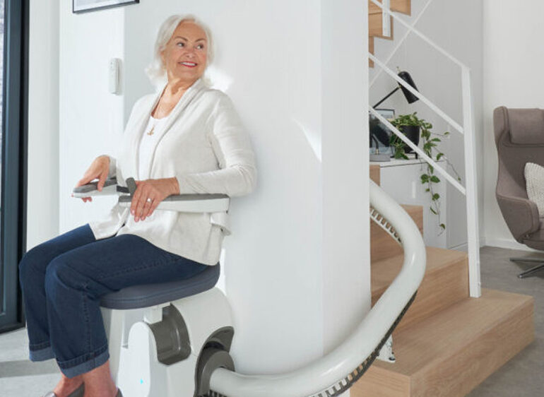 An older woman sits on a Flow X Curved Stair Lift at the base of a modern staircase in a bright, minimalist home, smiling as she prepares to ascend the stairs.