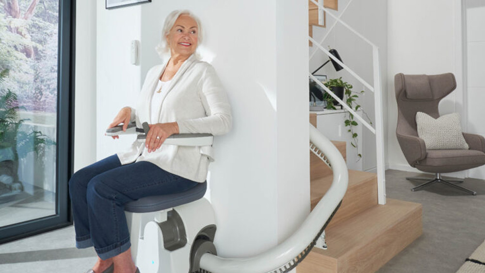 An older woman sits on a Flow X Curved Stair Lift at the base of a modern staircase in a bright, minimalist home, smiling as she prepares to ascend the stairs.