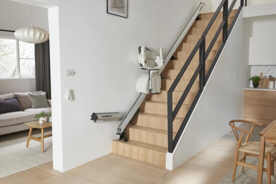 Straight stair lift with a hinged rail installed on a residential staircase, allowing the lower section of the track to fold upward and keep the hallway clear when the lift is not in use.