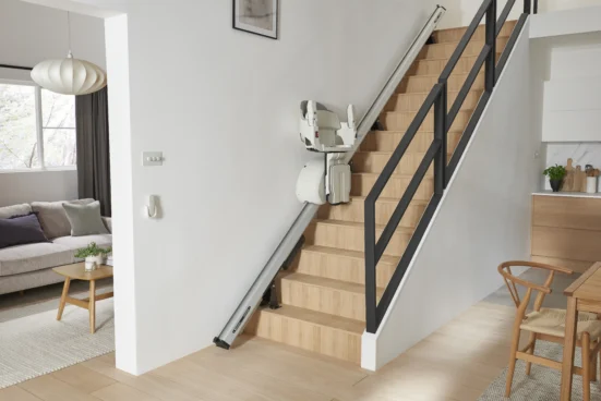Straight stair lift installed along a wooden staircase in a modern home, featuring a foldable seat, armrests, and footrest parked midway on the rail system to provide safe and convenient access between floors.