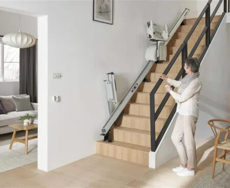 Woman standing beside an inclined platform lift installed on a residential staircase, demonstrating how the foldable wheelchair platform travels along a rail system to provide safe access between floors.