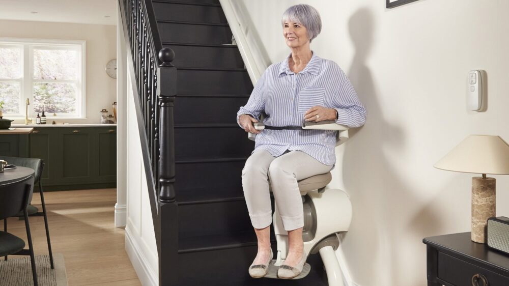 Woman using a stair lift to travel up a straight staircase in a residential home, featuring a comfortable seat, safety belt, and wall-mounted controls for improved accessibility.